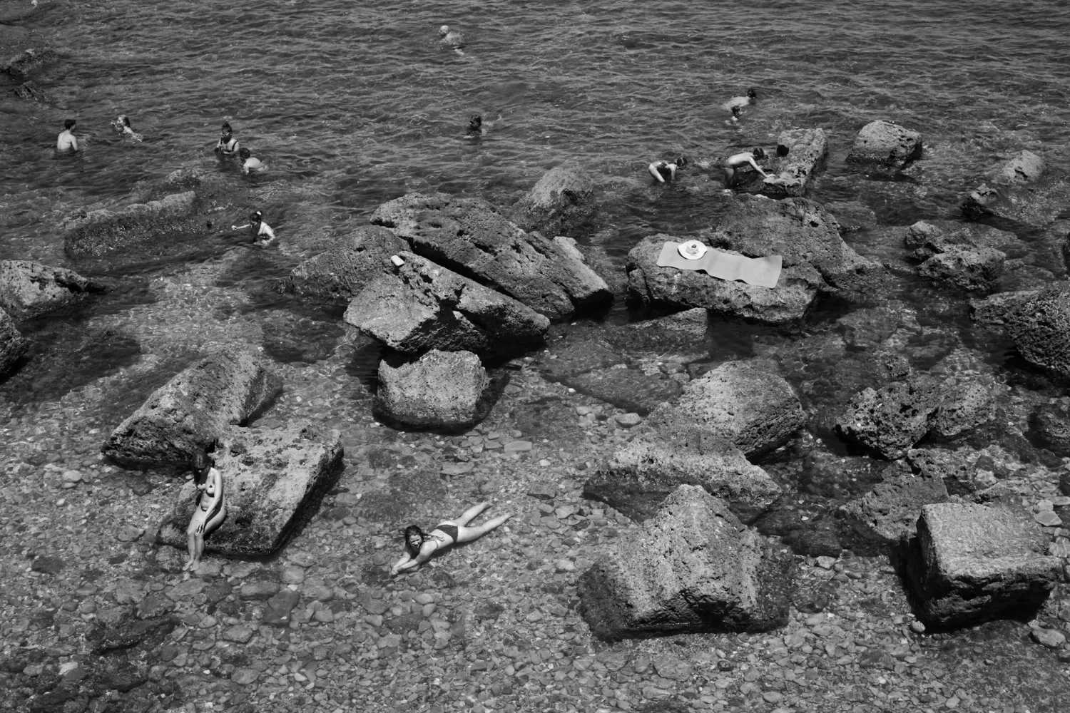 People relaxing at the beach, looking different direction, laying in the water with a lot of boulders/rocks surrounding them, almost makes them look like small ants.